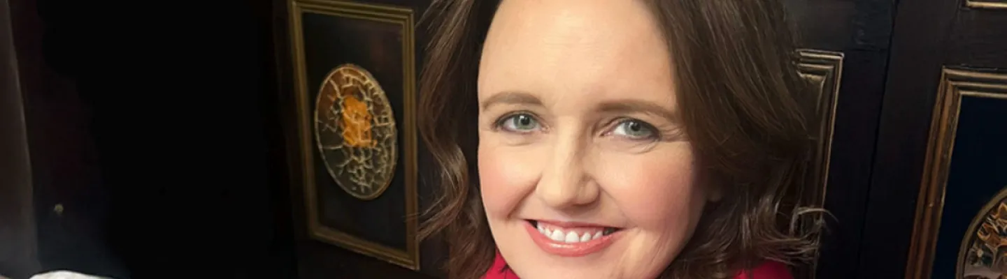 A woman is looking at the camera smiling. She is wearing a red jacket and sitting in front of a dark background with prints on the wall.