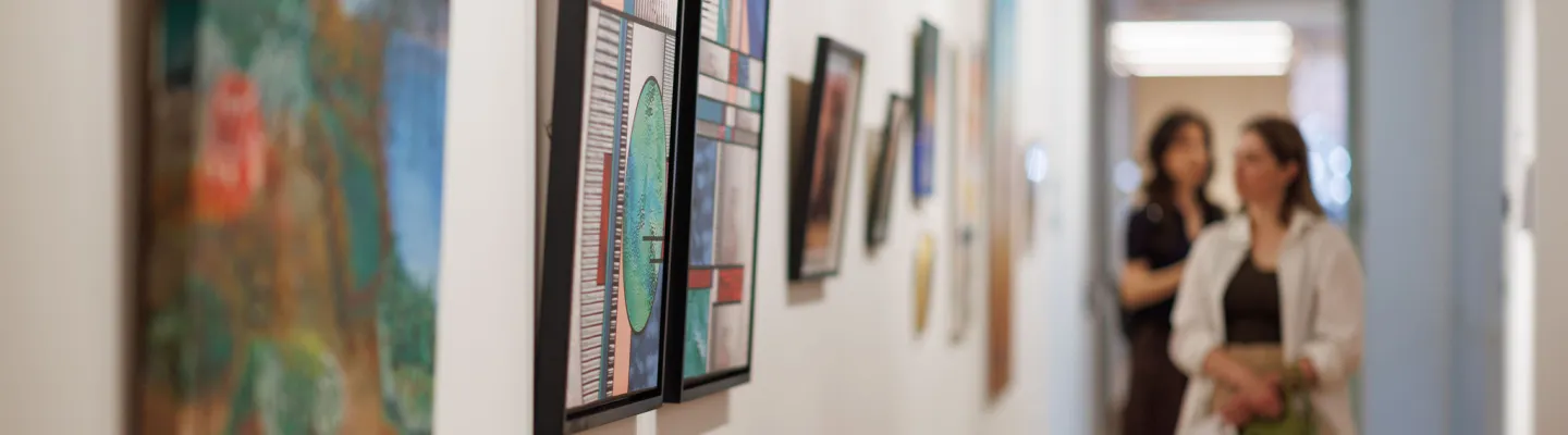 Long white gallery wall with a number of colourful artworks hanging and visitors looking. 