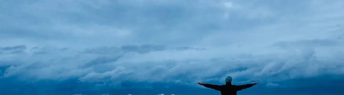 a man stands with arms spread wide on a beach in front of a large sky and surf