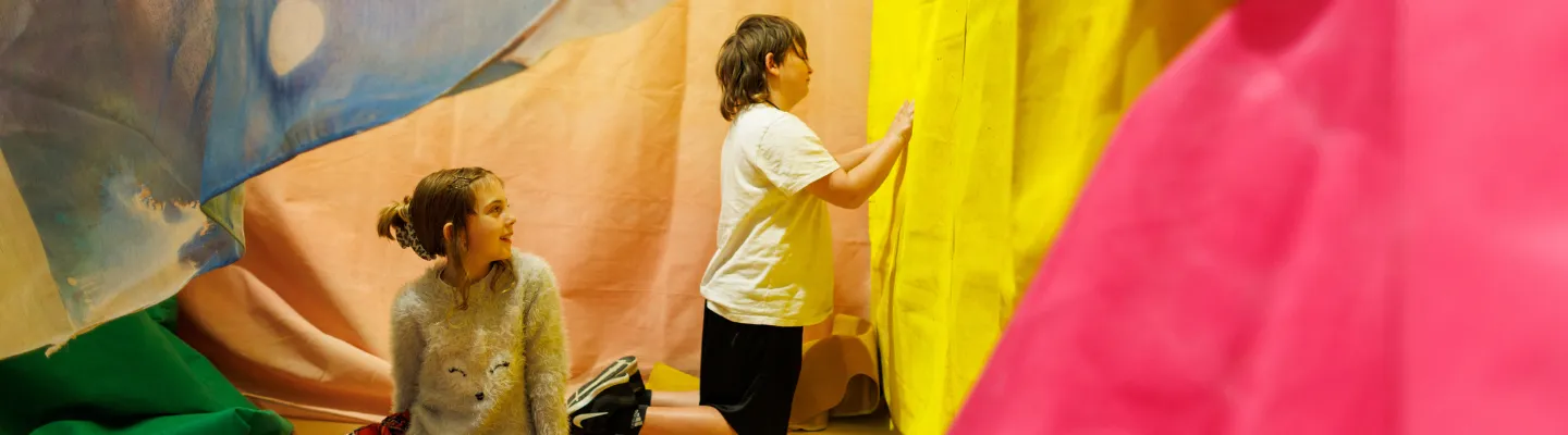 Two children are in a room with colourful fabric draped around them. One child is sitting on the floor wearing a sweater with an animal face design and patterned leggings, while the other child is kneeling and touching a bright yellow fabric wall. The room has wooden flooring and various colours of fabric hanging from above.
