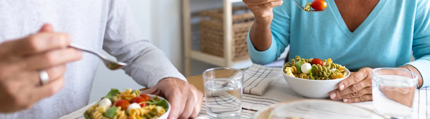 two people sitting at a table with bowls of pasta salad loaded with tomato and leafy greens.