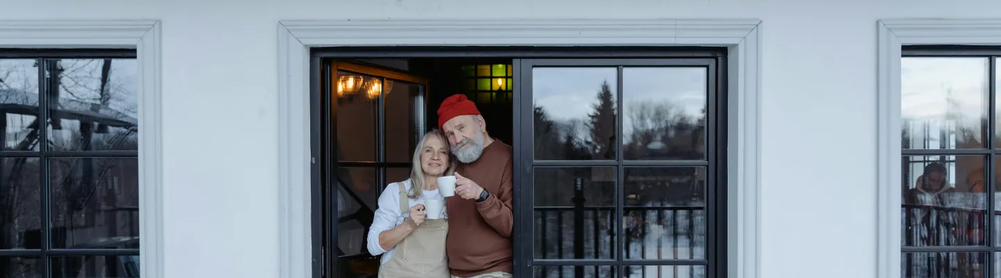 A woman and man stand in the doorway of a house. Both are holding white cups in their hands.