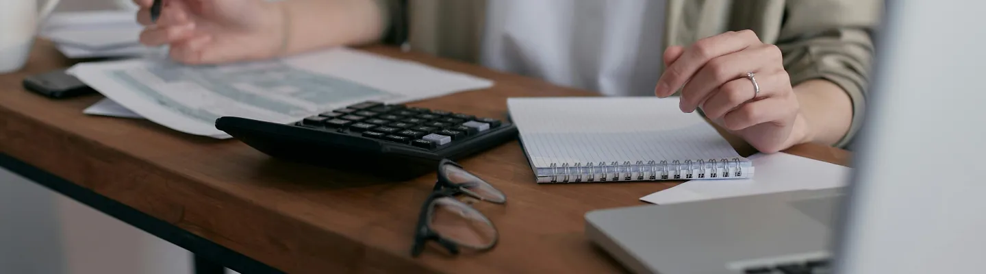 A person sitting at a desk, holding a pen. On the desk is a calculator, computer, glasses, notepad and some paperwork.