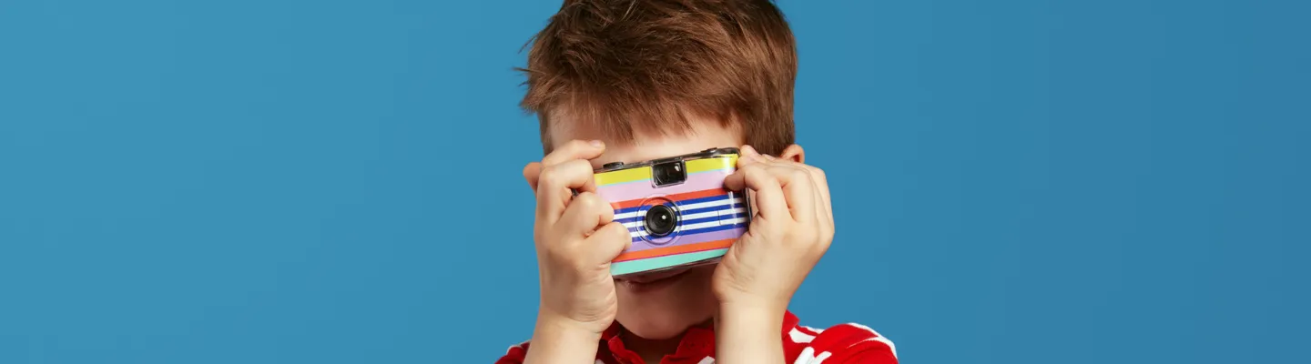 child holding up a rainbow coloured disposable camera up to their eye to take a photograph