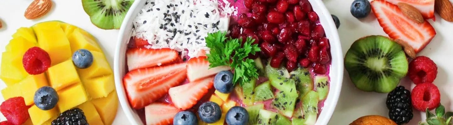 A colourful bowl of assorted fruits and yoghurt is surrounded by other fruits and nuts.