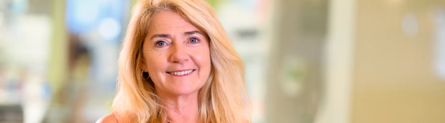 A woman with long blond hair is looking at the camera smiling. She is wearing an orange sleeveless top.