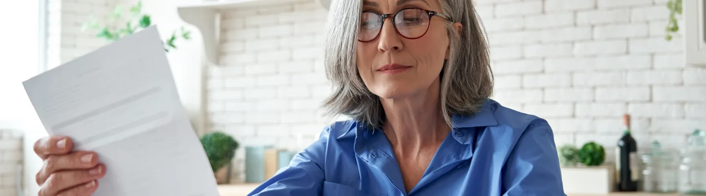 Woman at her kitchen table, looks at a bill. She has a calculator and a cup of tea.