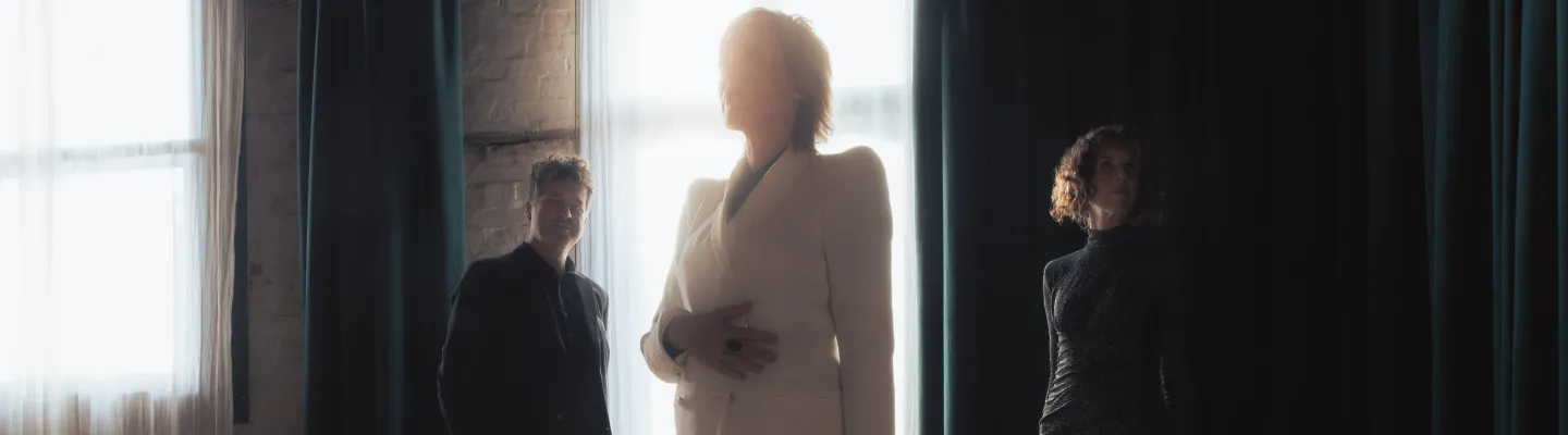 three people standing apart in an empty room with light shining through the curtains