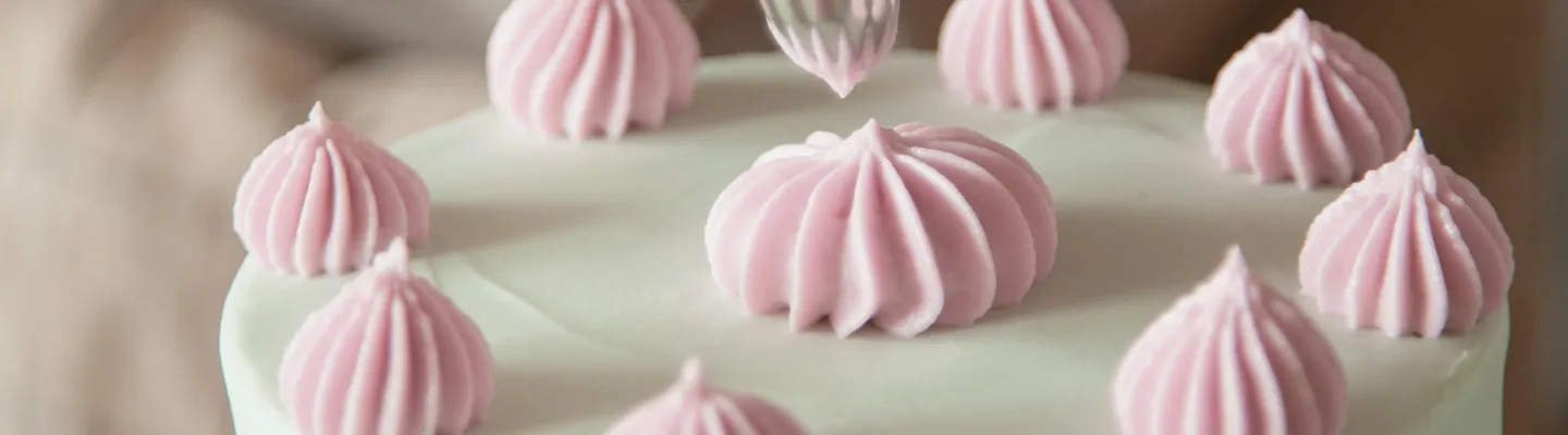 A person holding a piping bag is icing a white cake with pink icing flowers