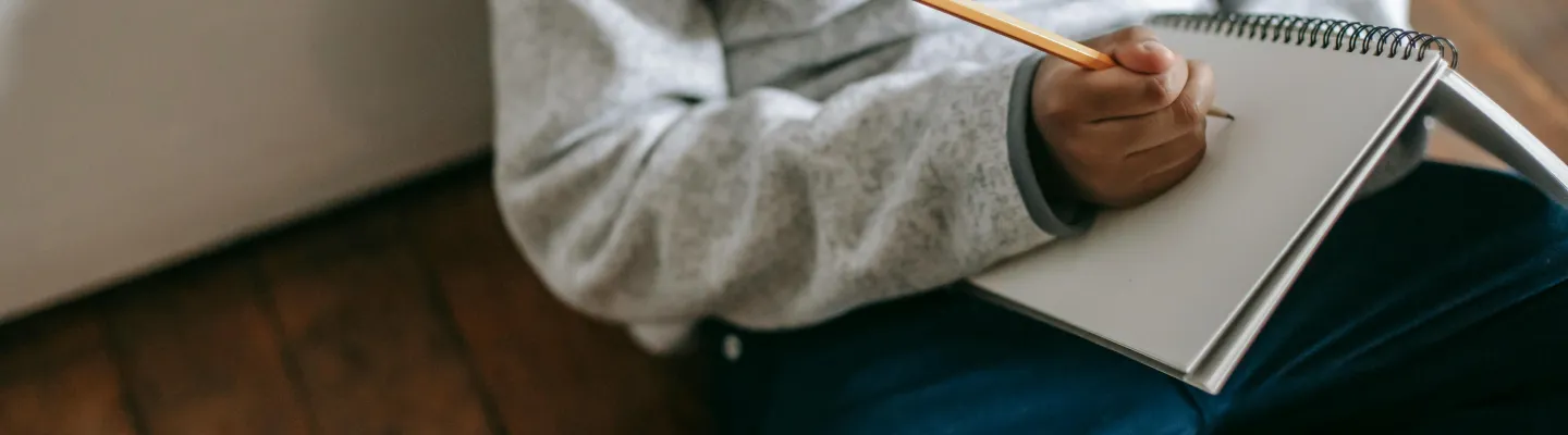 Child sits on floor holding a pencil and drawing on a notepad.