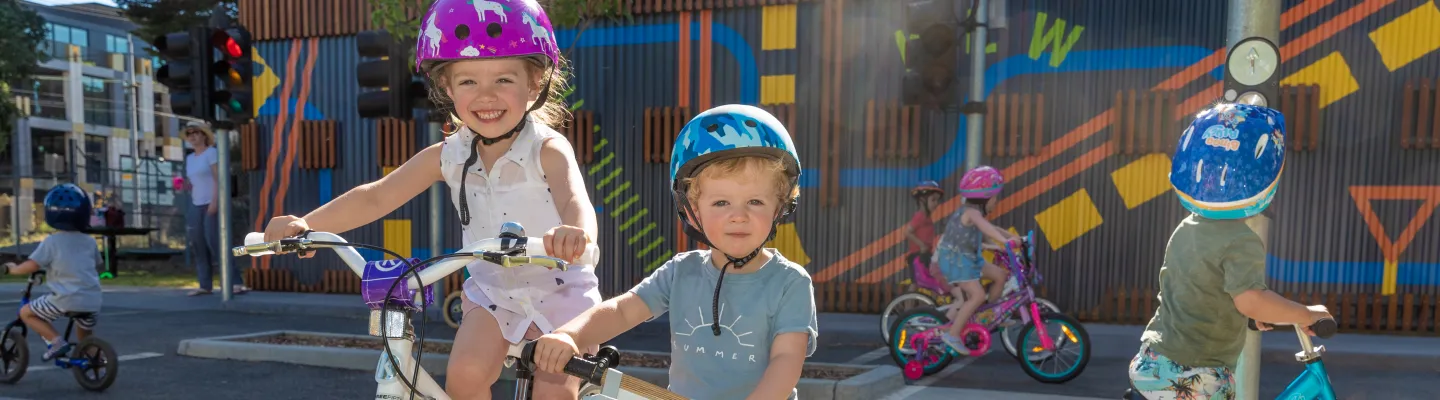 two children sitting on bikes. Lots of other children are riding around in the background. all are wearing bike helmets.