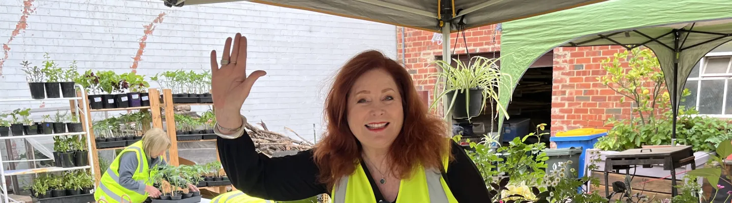 person standing in front of a plant stall at a market, smiling and waving. They are wearing a fluro yellow vest that says 'Boroondara Hard Rubbish Rehome'