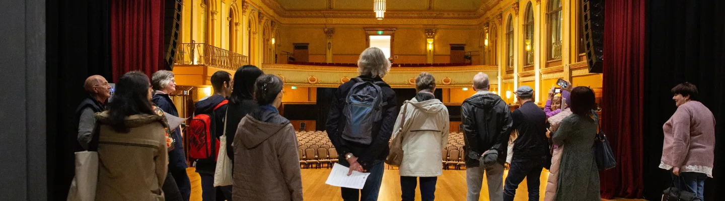 Group of visitors standing on a stage looking out to a large and grand hall