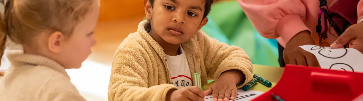 Two young children sitting at a table colouring in activity sheets with crayons