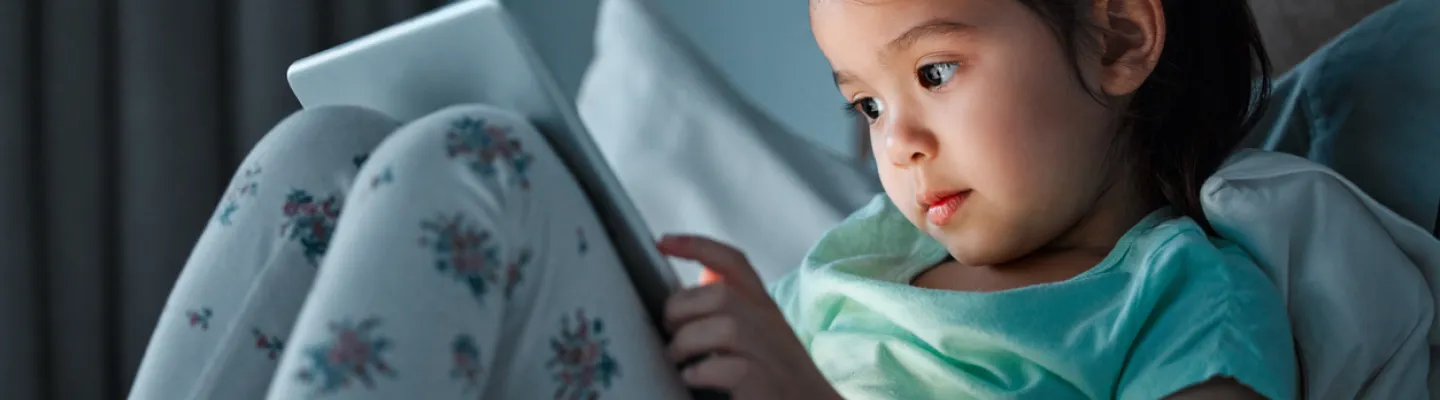 Young child sitting on a bed holding a tablet. Their face is illuminated by the screen.