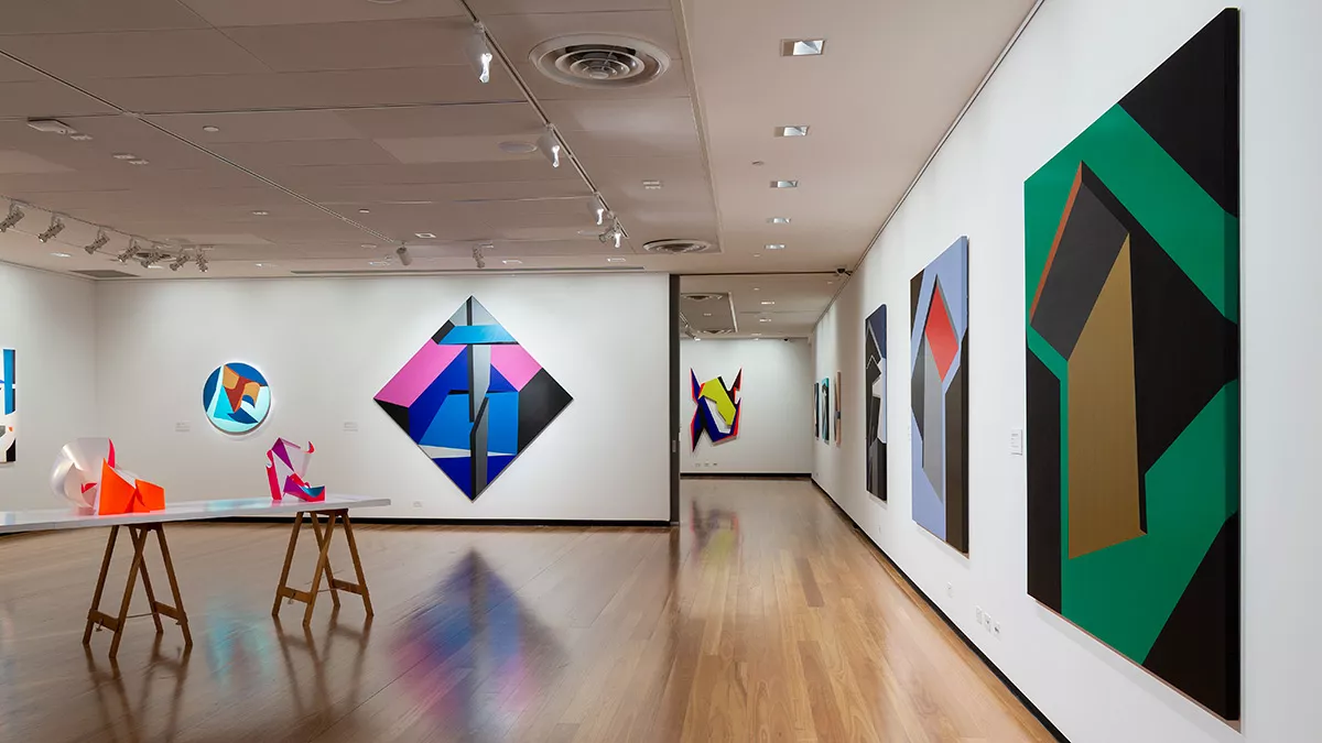 Installation view of 'Light Gestures' at Town Hall Gallery. Abstract geometric painting on the wall that are reminiscent of buildings or structures. Abstract sculptures on a table, made up of large flat surfaces and lots of angles.