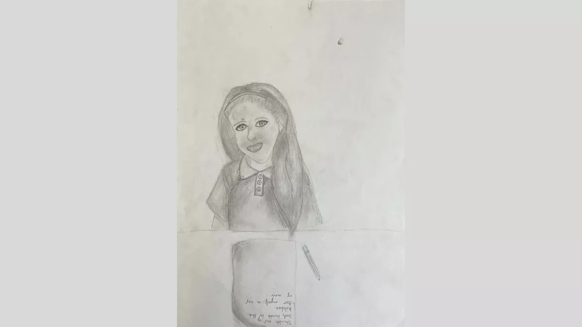 Graphite drawing of a girl sitting at a desk. She has a pencil and paper in front of her. She has long loose hair, is wearing a headband and is smiling.