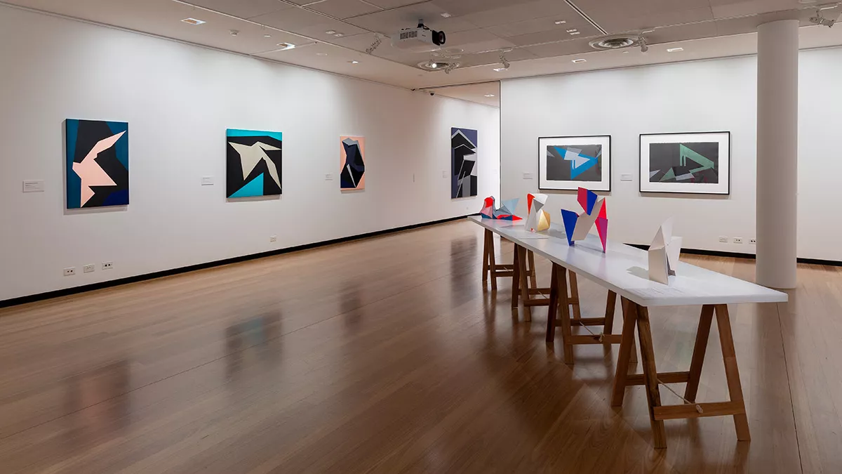 Installation view of 'Light Gestures' at Town Hall Gallery. Abstract geometric painting on the wall that are reminiscent of buildings or structures. Abstract sculptures on a table, made up of large flat surfaces and lots of angles.