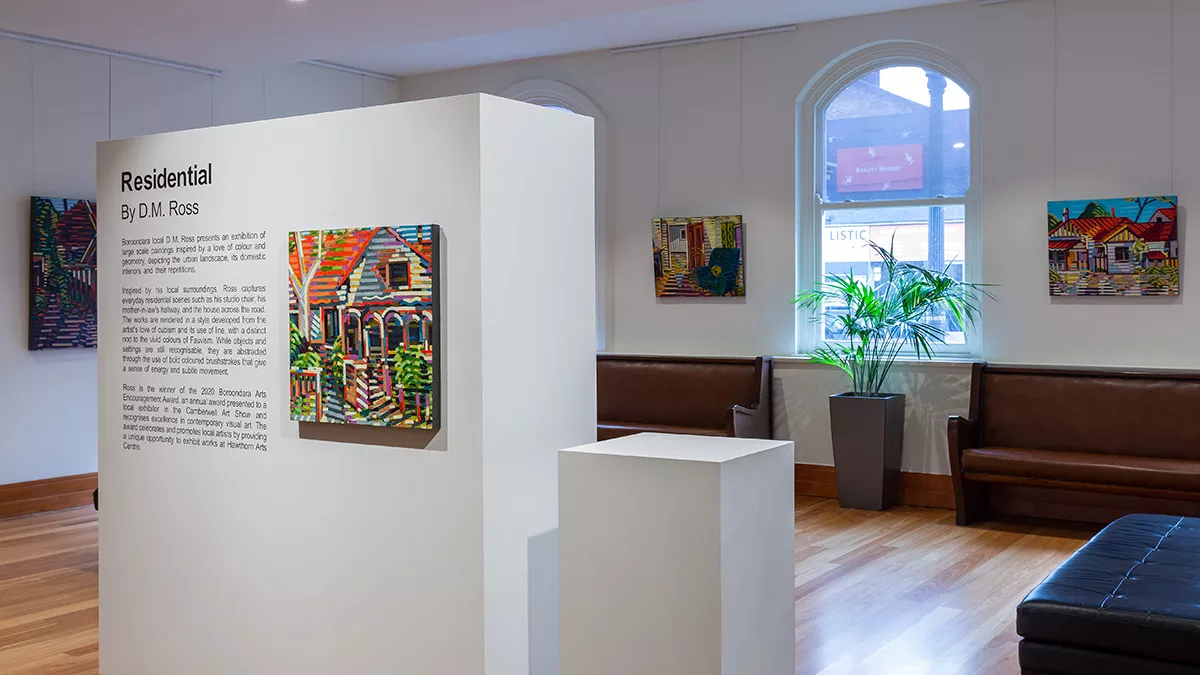 A section of movable wall in the centre of the exhibition space. On it is a blurb about the exhibition and a painting of the exterior of a house. Behind the wall are seats, windows and more paintings on the other walls.