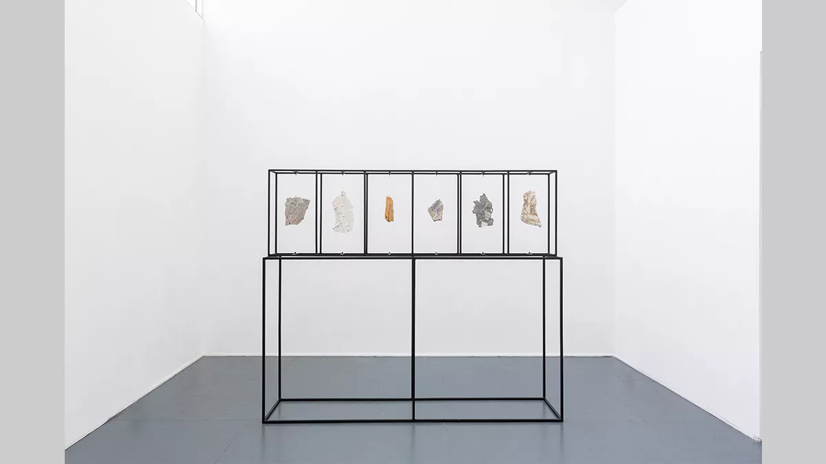 A rectangular steel structure in the centre of a plain space. At the top of the structure are what seem to be 6 smaller sections, in which are hanging abstract forms.