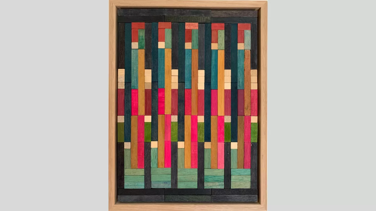 A framed piece of art made of wooden rectanges arranged in columns with bright pink, blue and some light and dark brown pieces in the middle of the columns surrounded by dark grey pieces