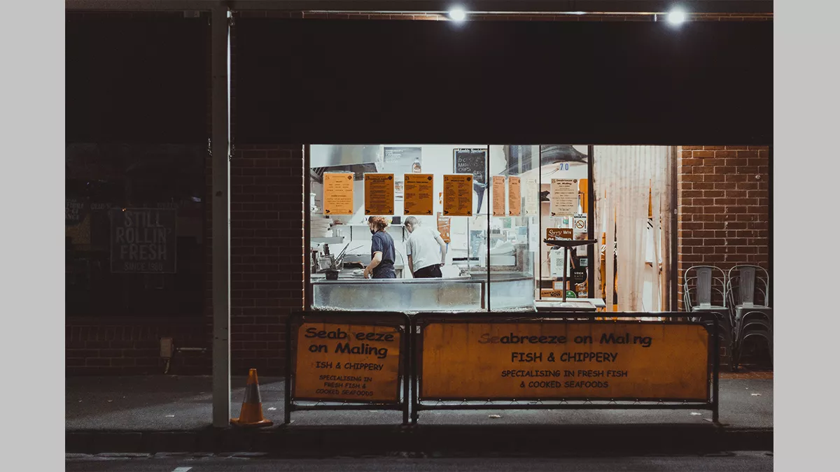 Photo by Mohan Fang of a bright shopfront at night through which you can see busy staff preparing fish and chips