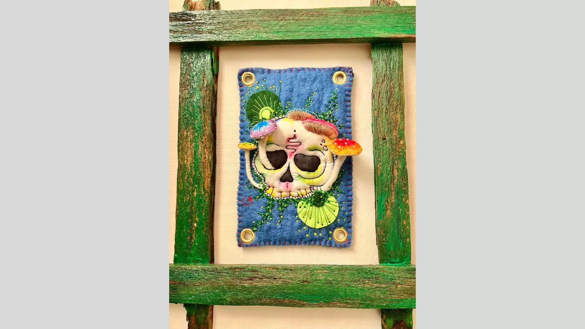A rustic green wooden frame with a piece of felt art in the middle of the frame showing a skull with mushrooms growing out of the skull which are raised from the felt so they emerge from the piece.