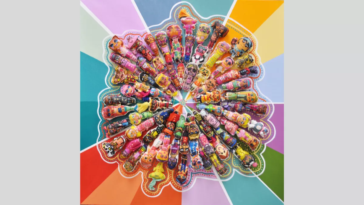 Artwork depicting plush toy dolls lying down in a circular formation, on a paint background of various colour panels.