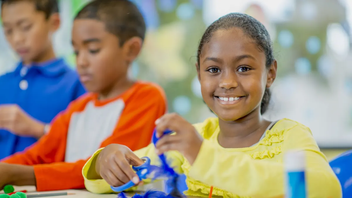 Three children participating in craft. Child in yellow shirt in focus, holding a pair of scissors cutting a feather.