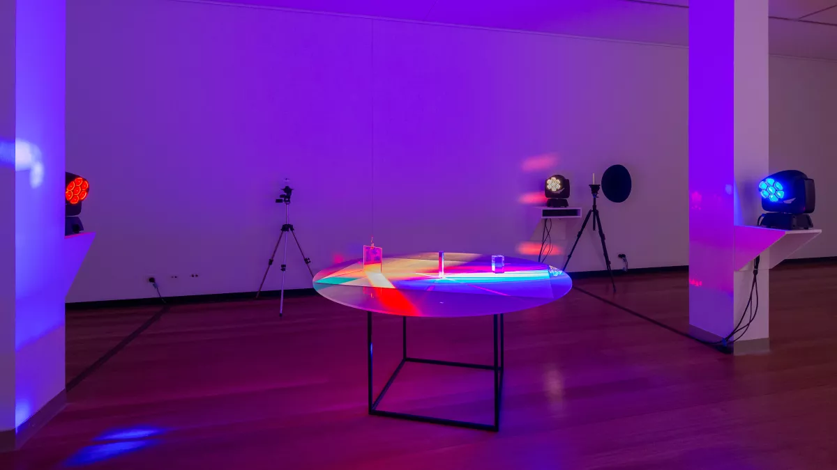 Art installation featuring three light projectors pointed at a round-top table, creating a mix of red, blue and yellow. There is a tripod near the far wall.