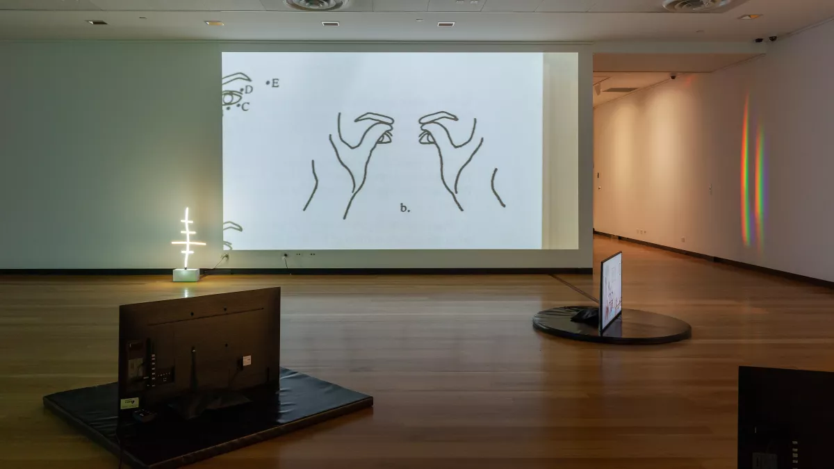 Indoor art installation space with hardwood floors and three scattered televisions. There is a drawing of thumbs pressed against eyes and eyebrows.