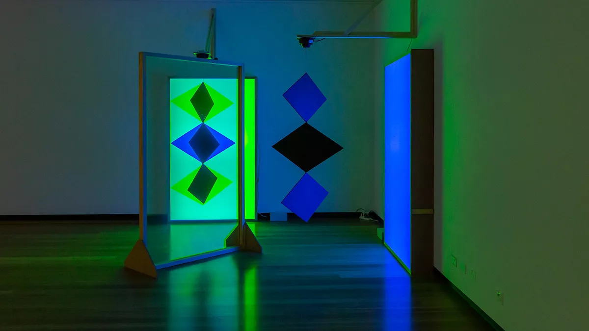 Art installation featuring three diamond shapes hanging from a fitting, and three diamond shapes on a glowing light green background.