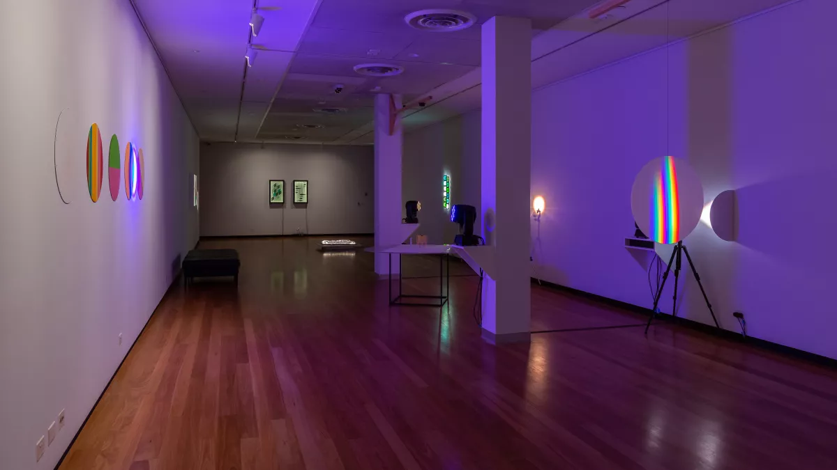 Long thin rectangular art installation space with two square columns and hardwood floors. There are multi-coloured artworks on round backgrounds to the left and right and two rectangular works in the distance.