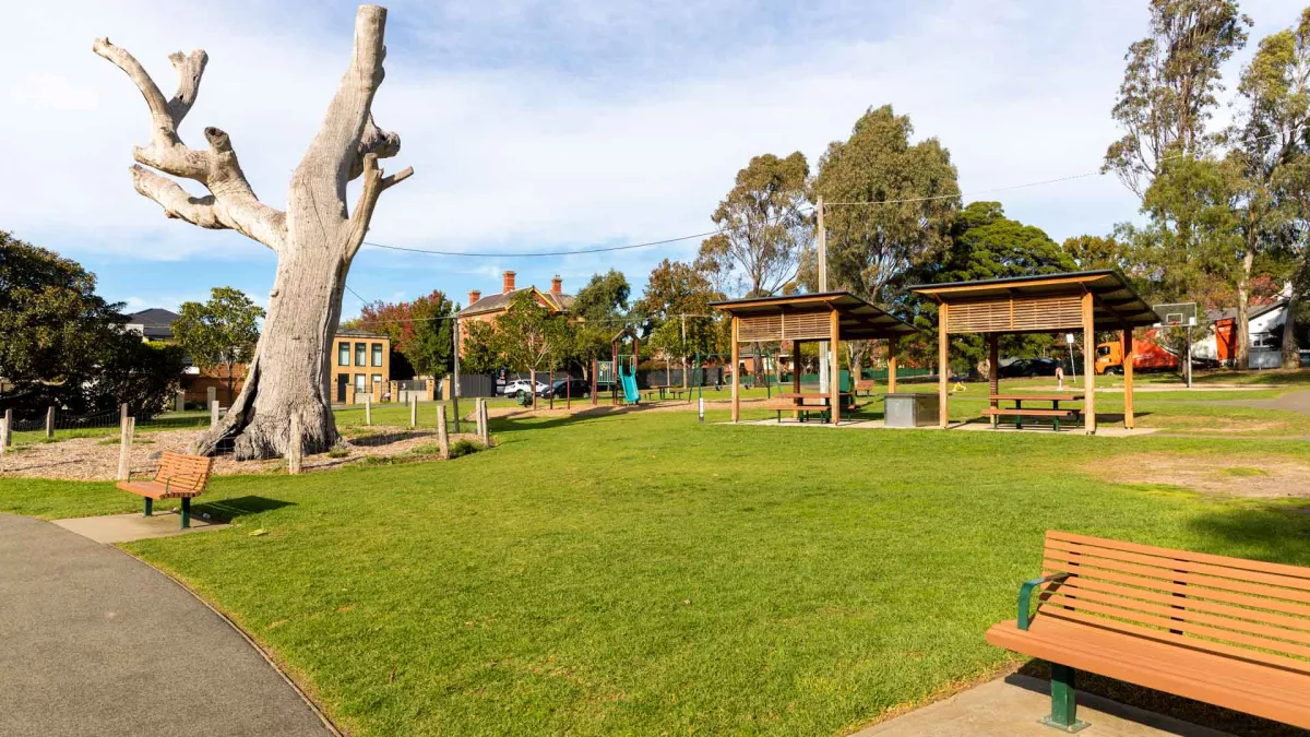 Grass area with a barbecue and two sets of tables and chairs with separate wooden shelters. There are two park benches and a footpath in the foreground and a large grey tree stump to the left. 