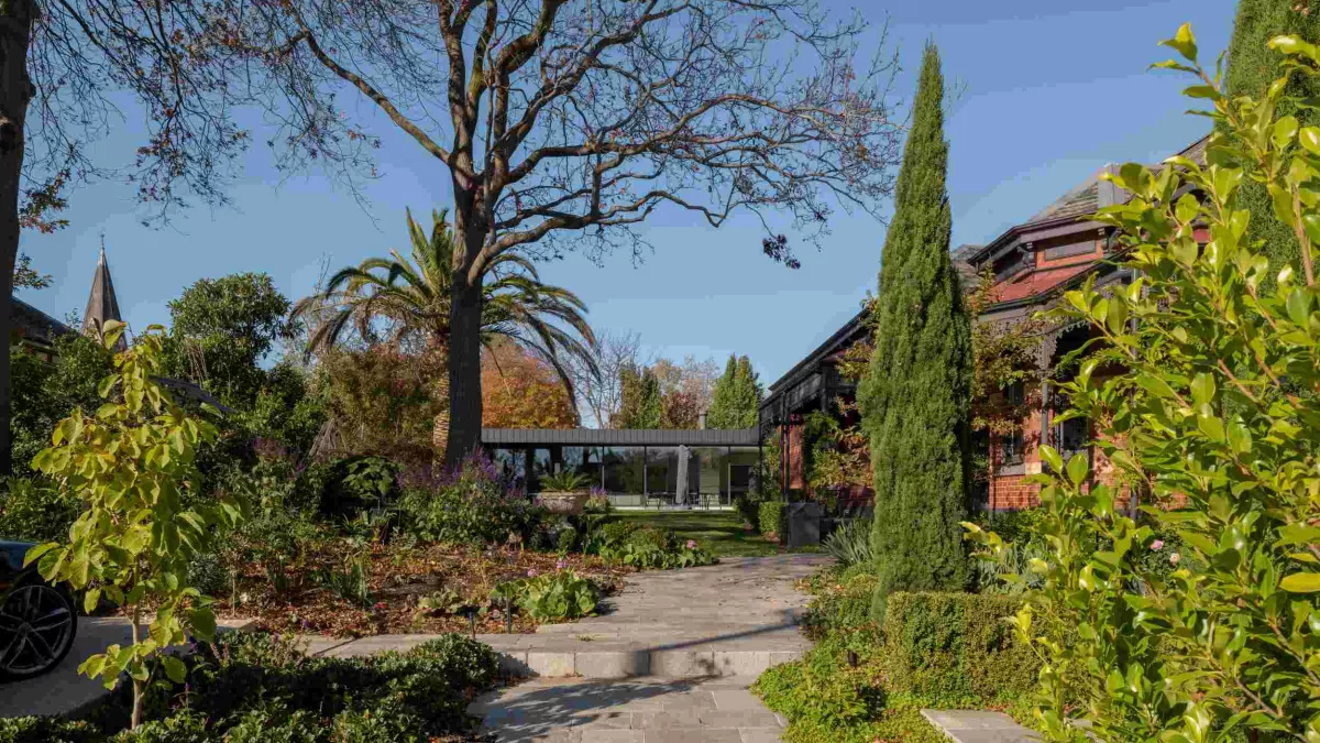 A glass-walled room extends from a heritage red-brick house in a landscaped garden