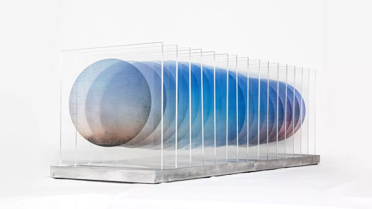 Sculpture artwork comprising 16 clear squares alongside one another on a perspex and aluminium base. Each square contains a single blue and green circles. The background is white.