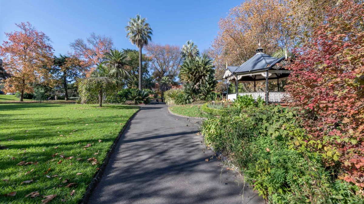 Concrete path with grass areas on either side. There is a covered rotunda with lattice patterns to the right, and tall trees in the distance.