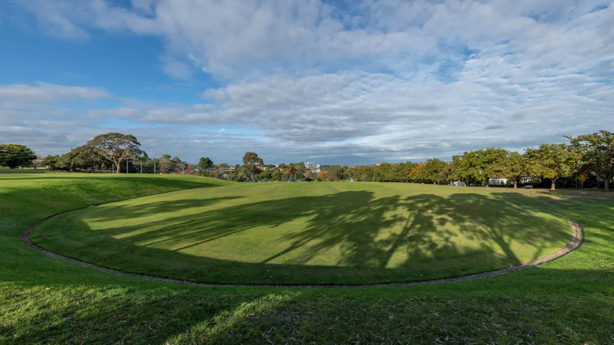 Oval-shaped grass field with a line of bricks around its boundary and sloped grass around its edge. 