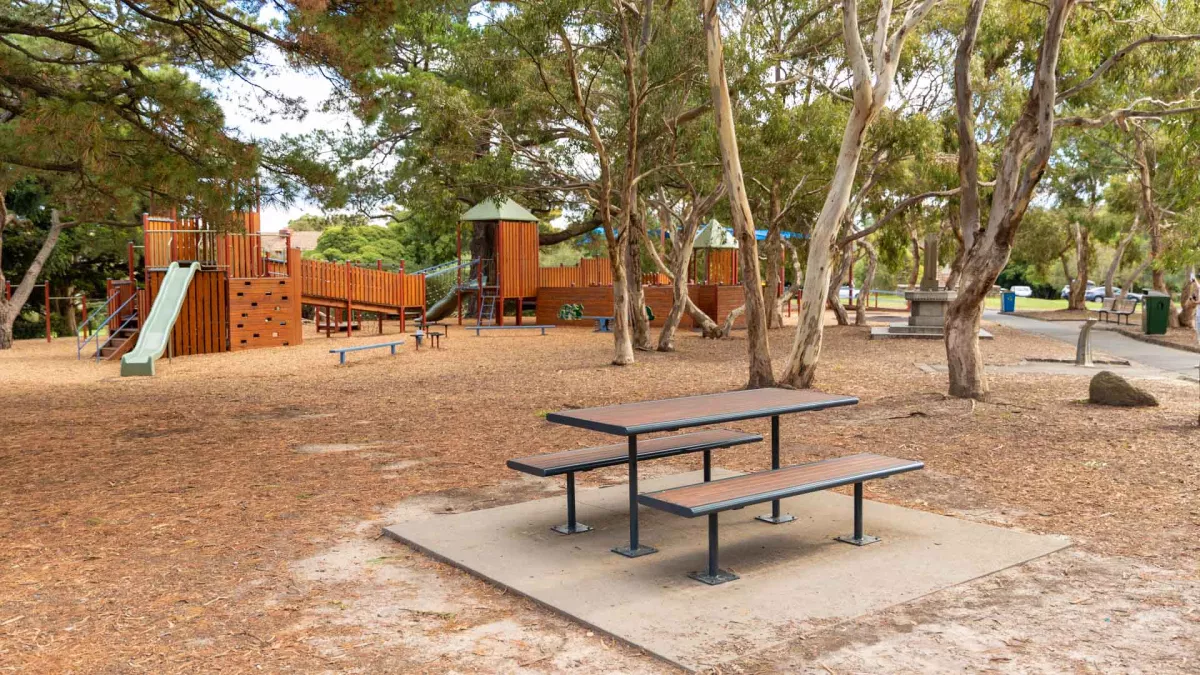 Table and two bench chairs fixed to ground with poles, with trees and a playground in the background.