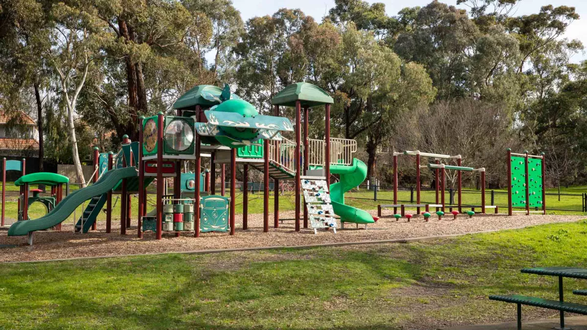 Playground featuring a red and green climbing structure with two slides, and two smaller structures. There is a table and chair set to the right and tall trees in the background.