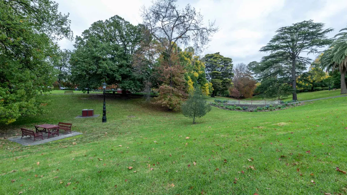 Large grass area which slopes into a small valley with a walking bridge, among mature trees. There is a light post and table with two bench seats to the left.