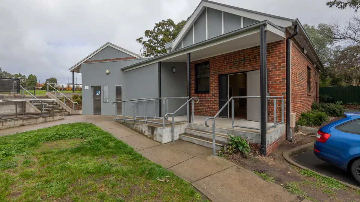 Single-storey brown brick building with triangular grey roof, with steps up to a sportsground to the left. There is a walking path and grass in the foreground.