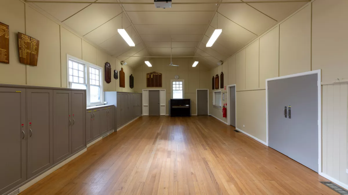 Inside of a hall with polished floorboards and a piano at the far end. There are two grey doors on the right and grey cupboards and a window along the left.