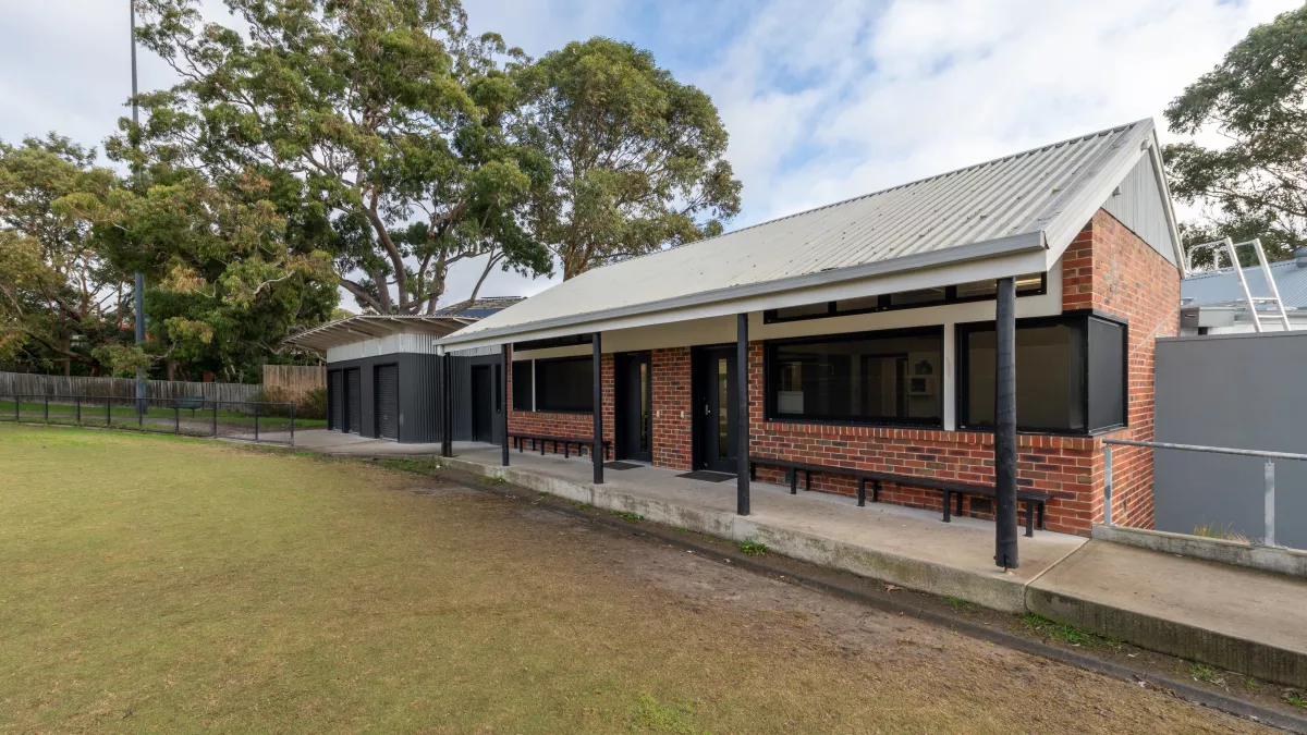 Single-storey brown brick building with triangular roof and black window frames. There are tall trees in the background.