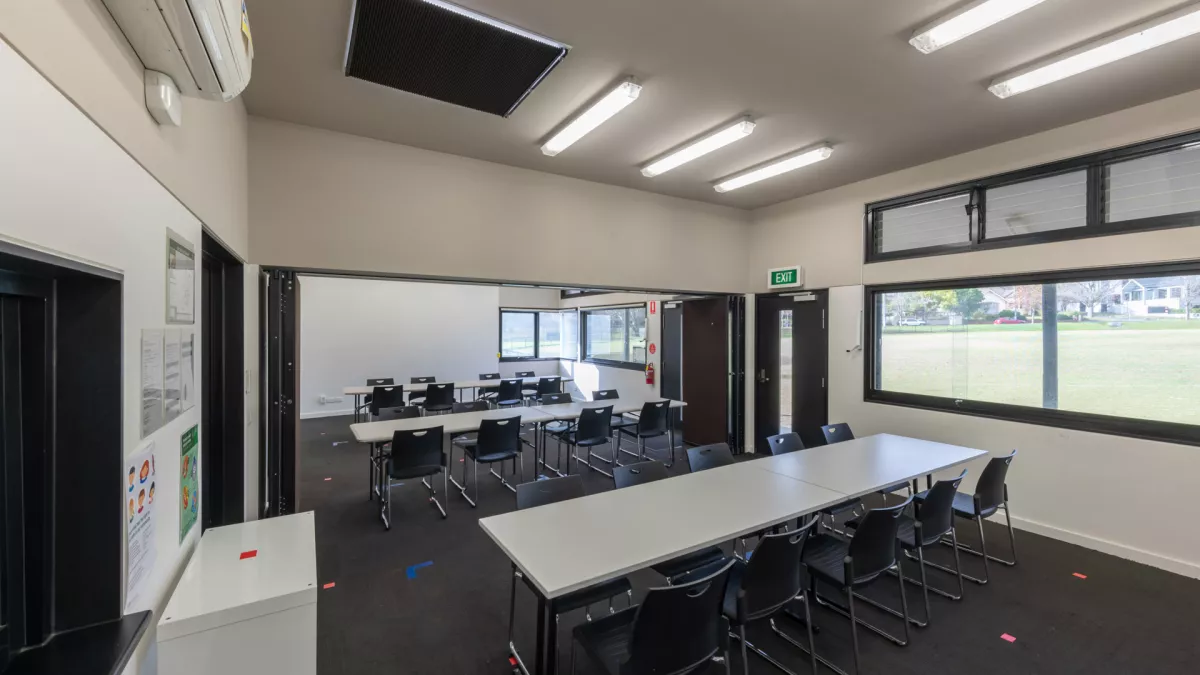 Room with three white tables and 30 black chairs. There are several wide windows on the right and a sportsground outside.