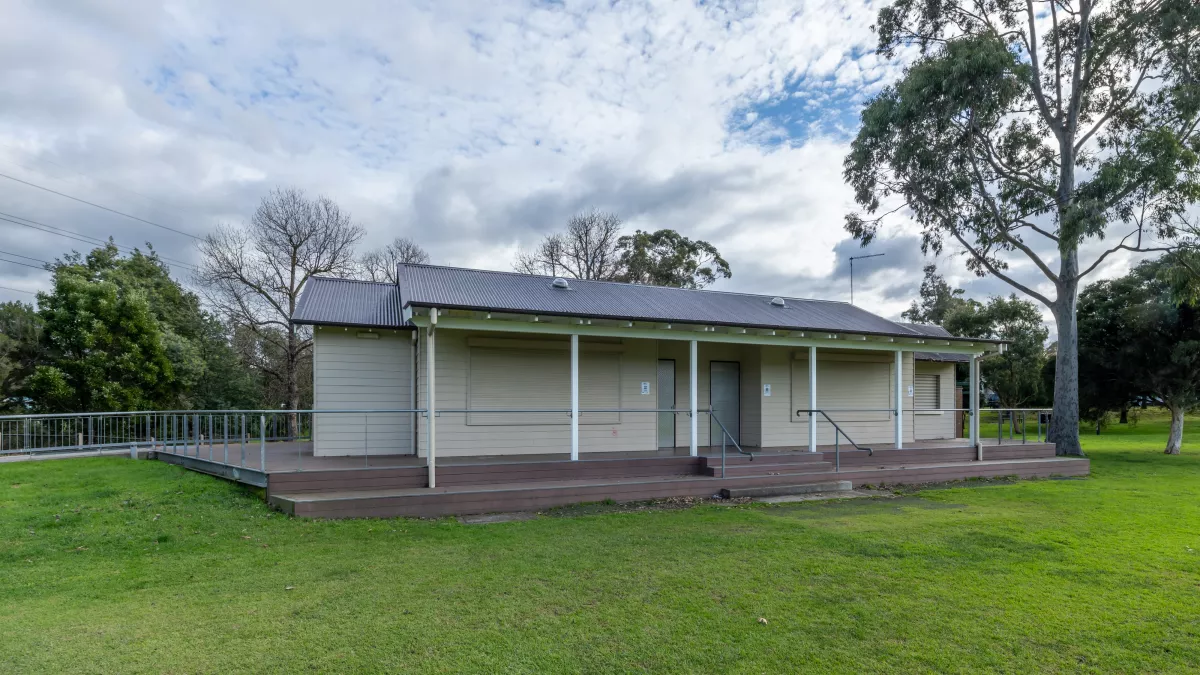 Single-storey cream white weatherboard building with sloped corrugated roof, on the edge of a sportsground. There is a tall tree to the right and small trees in the background.