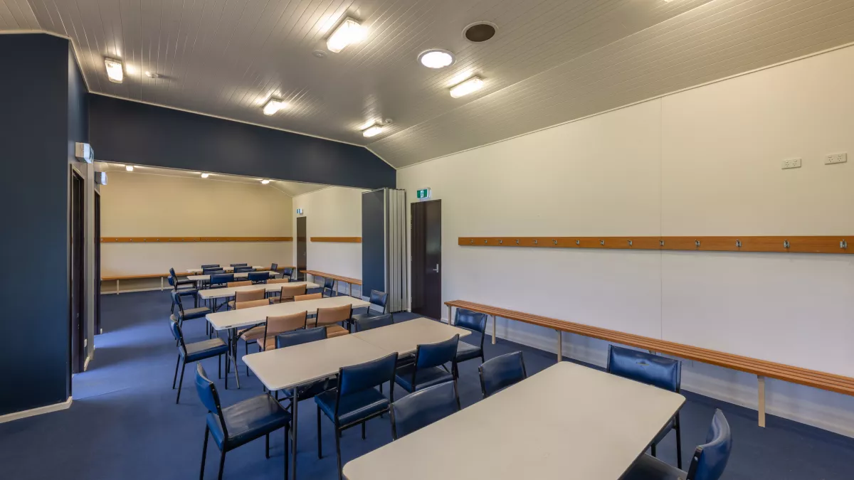 Long thin room with six white tables and 30 blue and brown chairs. There are coat racks and bench seats along the walls, and two doors to the left and right. The lights are switched on.