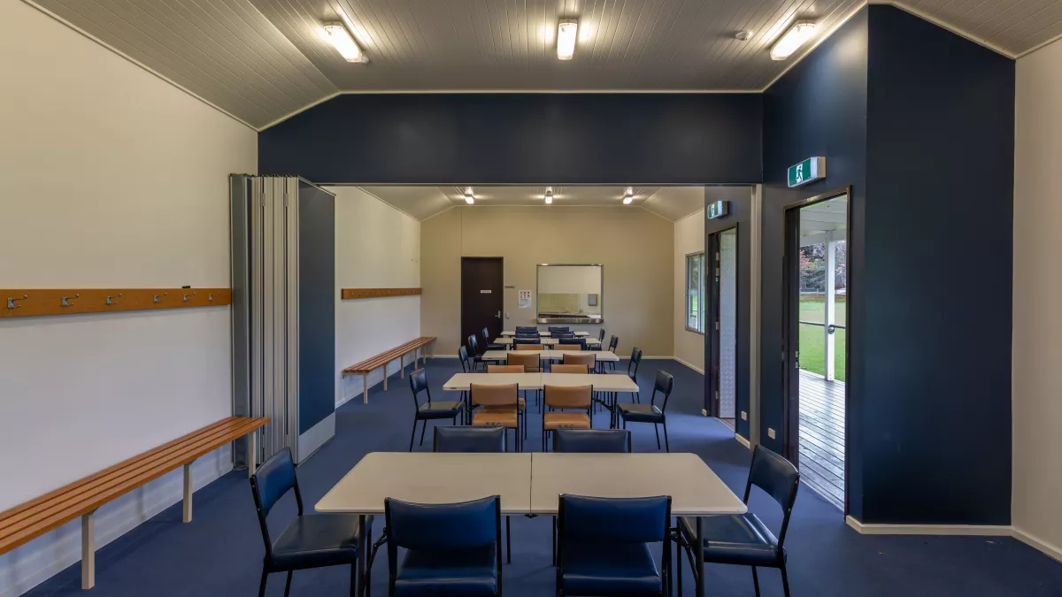 Long thin room with six white tables and 30 blue and brown chairs. There are coat racks and bench seats along the walls, two doors to right and a kitchen at the far end. The lights are switched on.