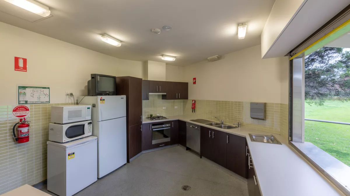 Kitchen with white benches, black cupboards and a grey floor. Items include a fridge, freezer, two microwaves and a fire extinguisher. Grass is visible through a window on the right.