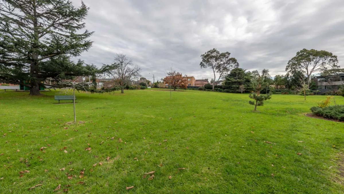 Large grass area containing several small trees. There are tall trees and houses in the distance and a park bench under a large tree to the left.